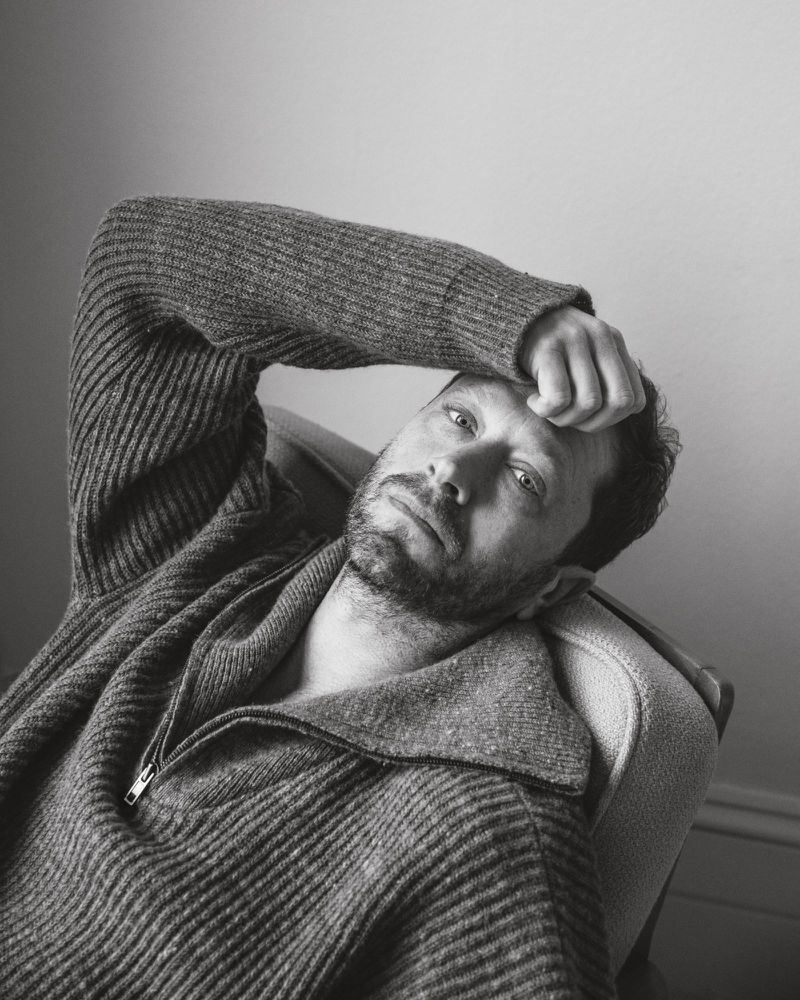 Portrait of adult man with beard and grey sweater, lying back on armchair. Indoors. Black and white photograph. Head and shoulders.