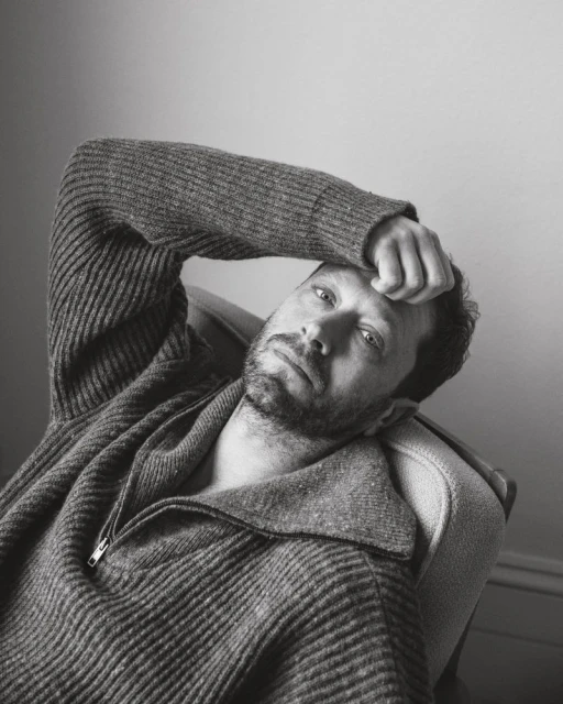 Portrait of adult man with beard and grey sweater, lying back on armchair. Indoors. Black and white photograph. Head and shoulders.