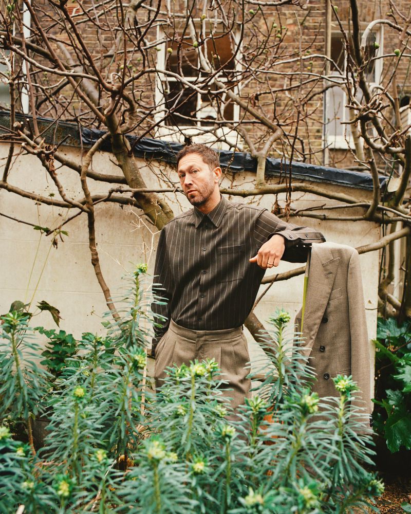 Adult man in striped shirt leans against a suit jacket in an outdoor garden setting with stone wall and bare branches in background.