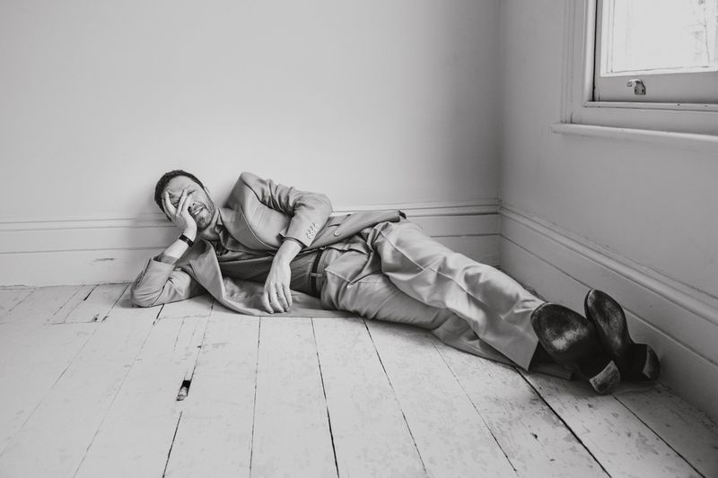 Man in a tan suit poses while lying on a wooden floor in front of a white wall and window, creating a studio fashion shot in black and white.