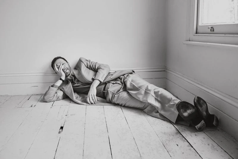 Man in a tan suit poses while lying on a wooden floor in front of a white wall and window, creating a studio fashion shot in black and white.