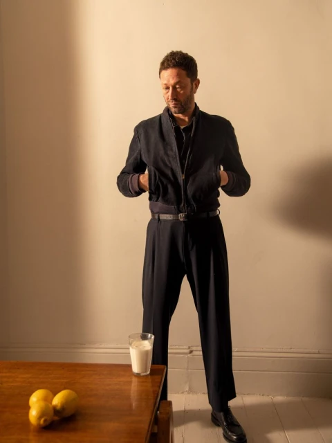Full-length shot of a man in a dark blue jacket and pants standing indoors, with a glass of milk and lemons on a wooden table.