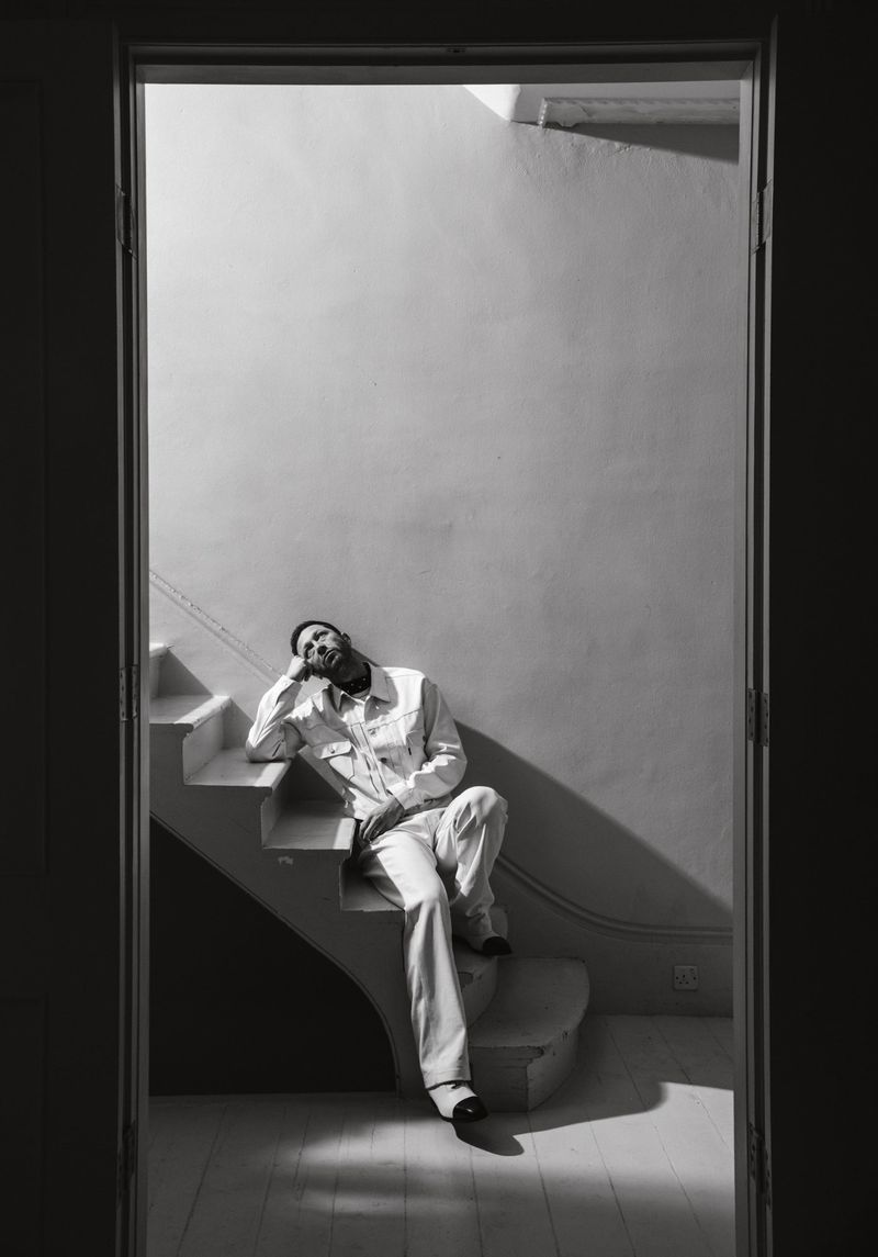 Monochrome portrait of a bearded man in a white suit sitting on white stairs, framed by a doorway in soft, natural light.