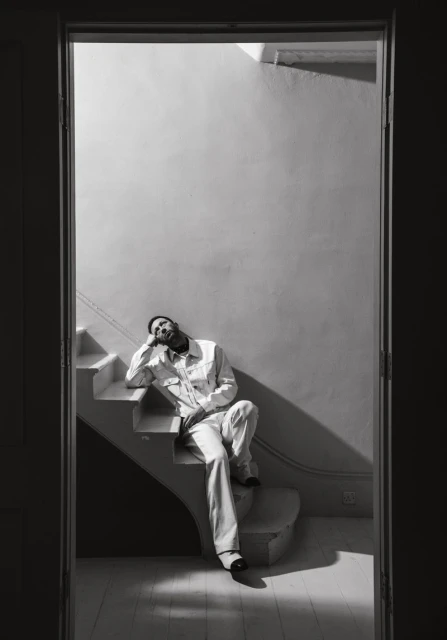 Monochrome portrait of a bearded man in a white suit sitting on white stairs, framed by a doorway in soft, natural light.
