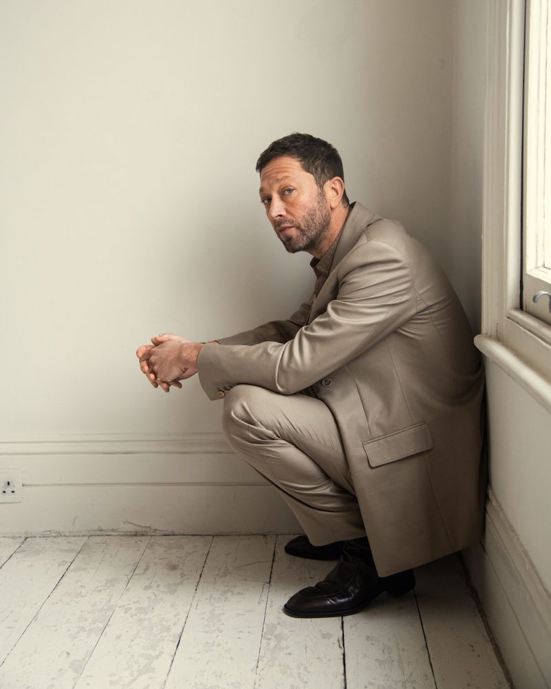 Mature man in suit crouching indoors looking at camera in portrait style. Neutral studio background with white walls and wooden floors.
