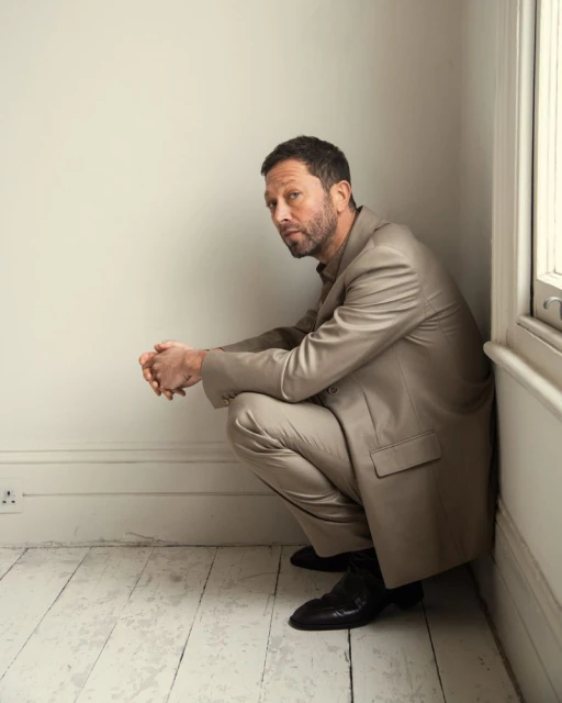 Mature man in suit crouching indoors looking at camera in portrait style. Neutral studio background with white walls and wooden floors.