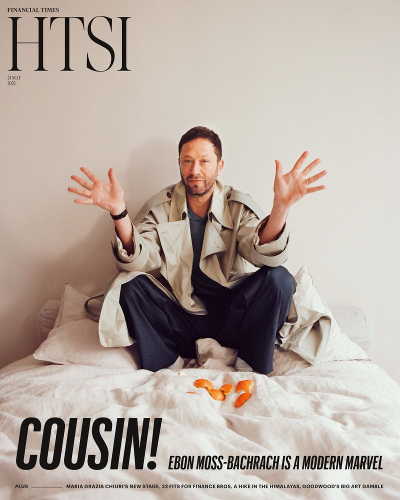Magazine cover featuring Ebon Moss-Bachrach in a beige trench coat, seated on a bed, with outstretched arms and a neutral facial expression.