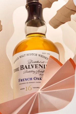 Close-up of a Balvenie 16 Year Old Single Malt Scotch Whisky bottle with paper art, studio setting, and warm lighting.