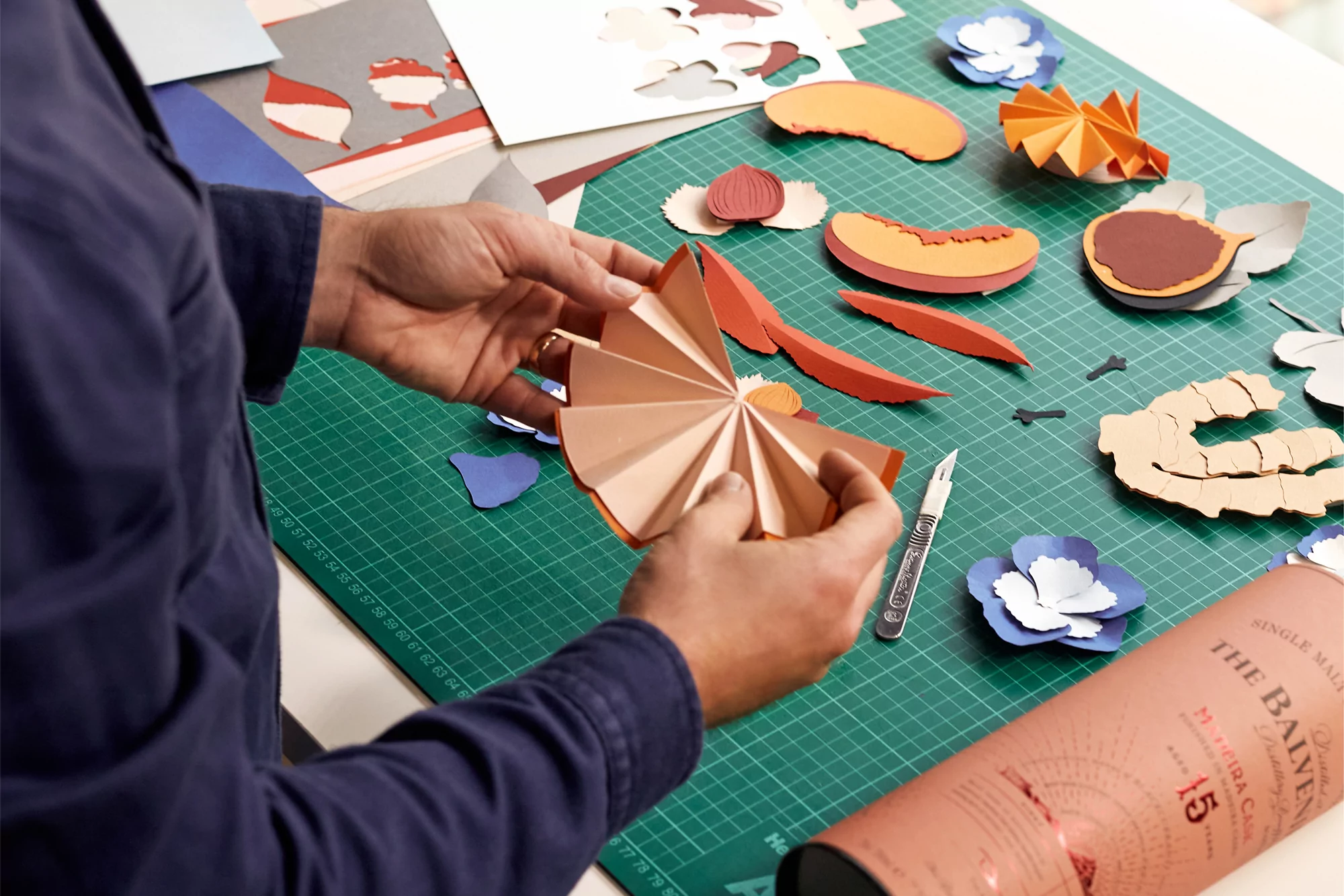 Hands fold peach paper, arranged on green cutting mat with paper flowers, tools, Balvenie whisky bottle. Table top paper craft flat lay.