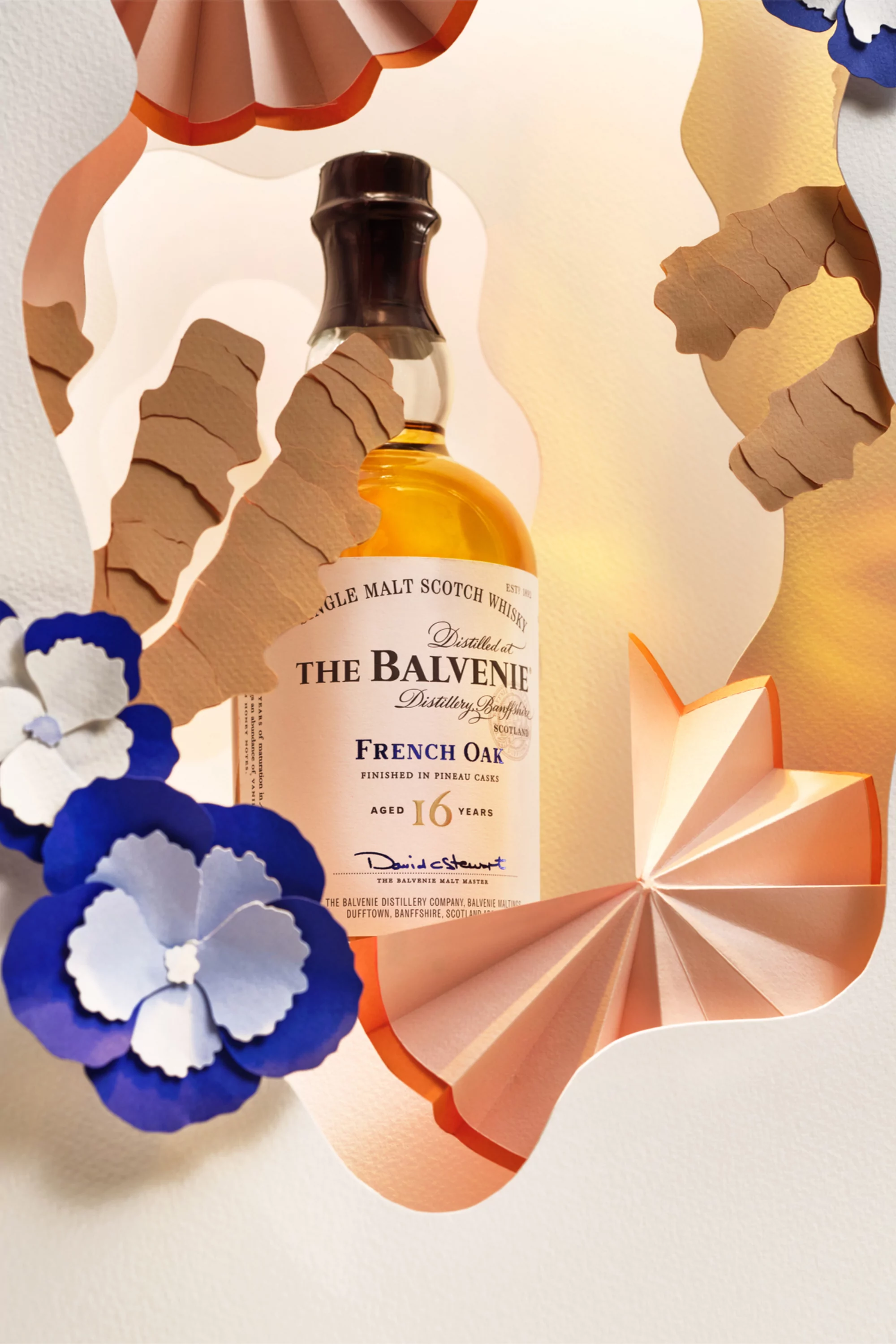 Bottle of Balvenie Scotch whisky surrounded by a paper art display with blue and orange paper flowers and other decorative shapes.
