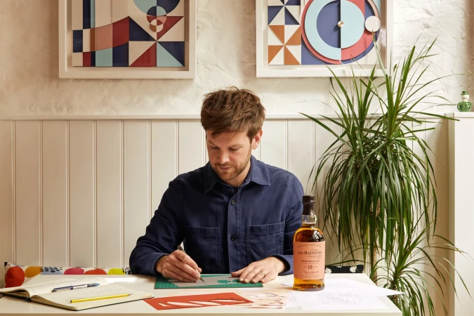 A man is seated at a table working on an art project using a stencil, knife, and paper against a backdrop of geometric art and houseplants.