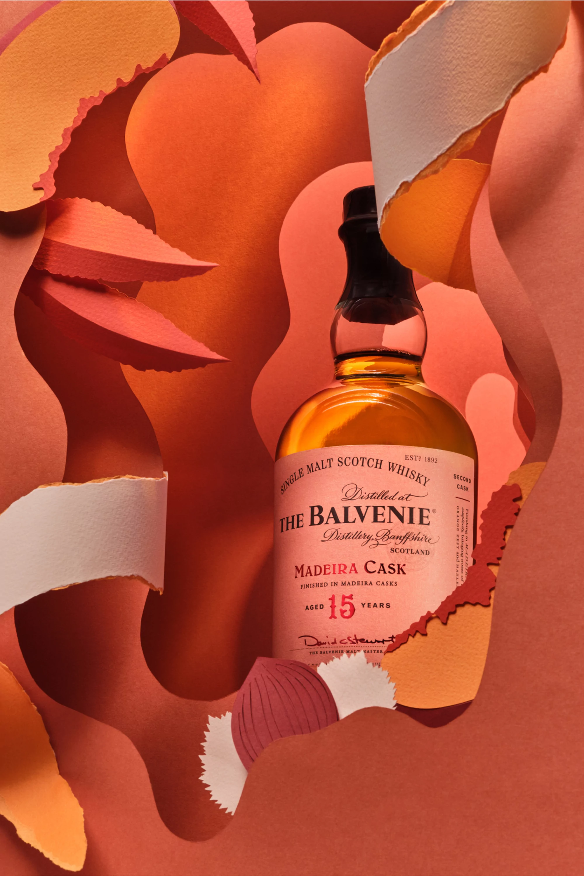 Balvenie Scotch whisky bottle in artful still life, featuring paper-cut background and product close-up with amber liquid.