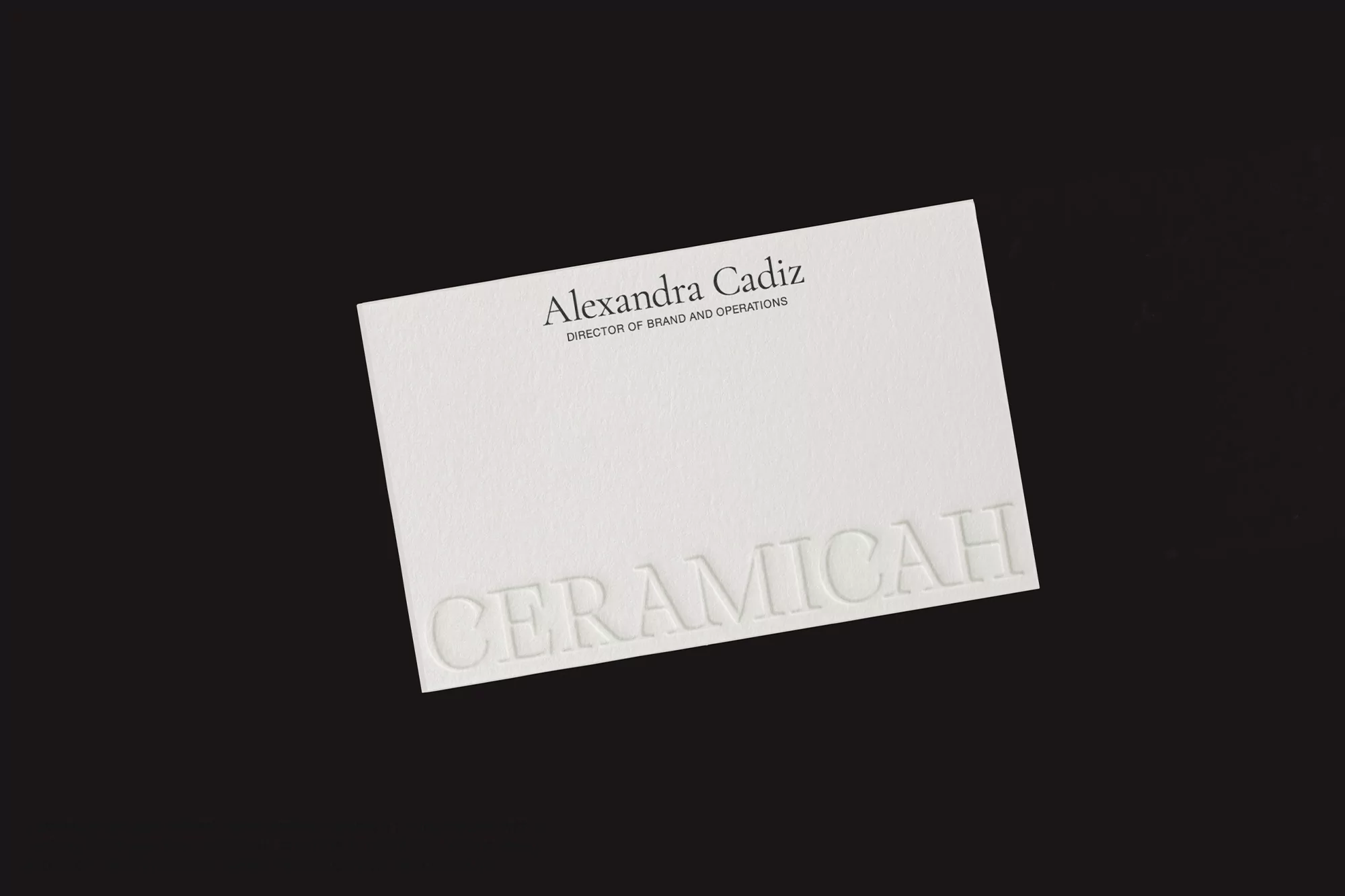Close-up of a white business card on a black background with the name Alexandra Cadiz and the word CERAMICAH embossed on it.