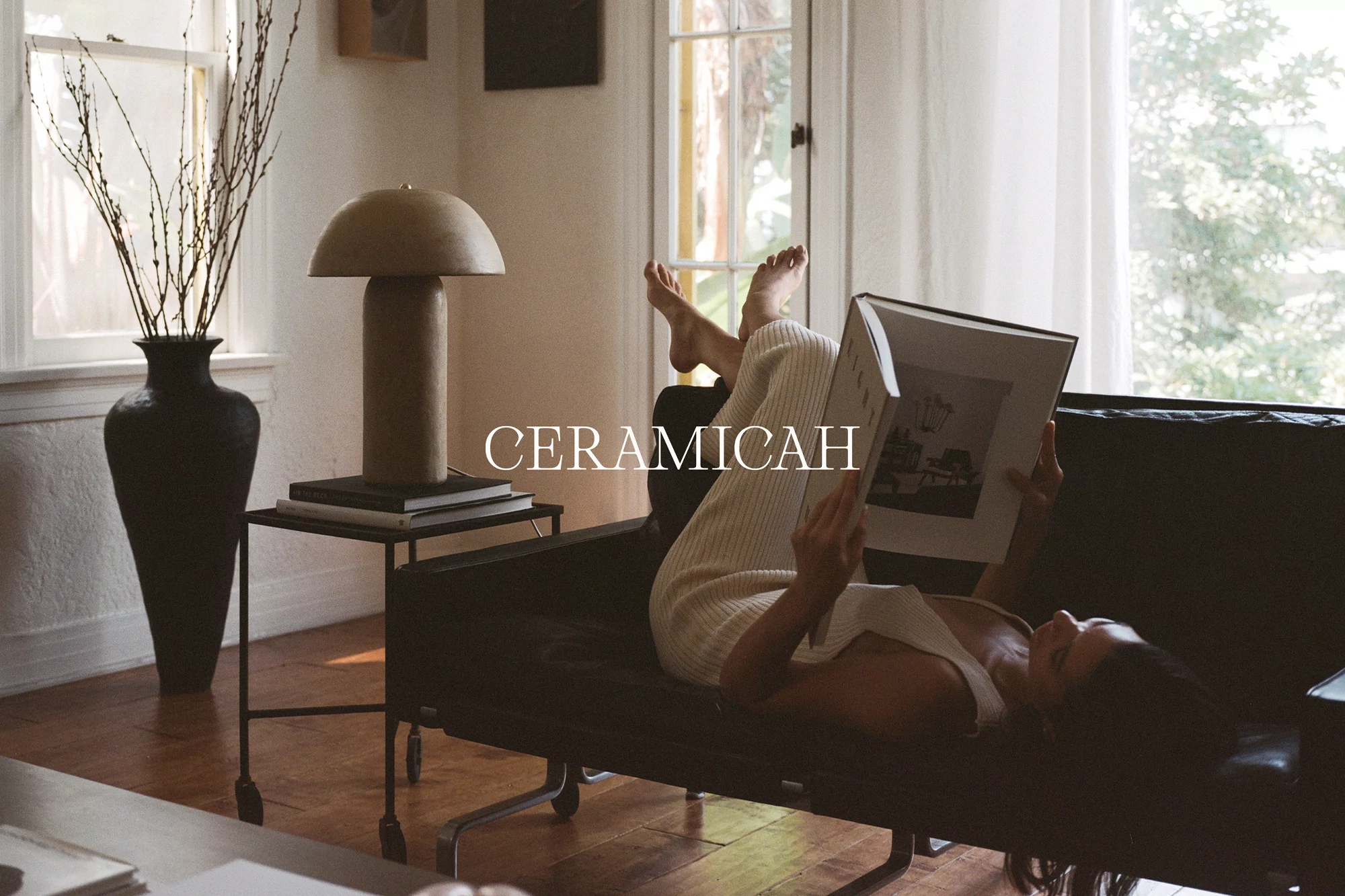 Woman reading a book while laying on a black couch indoors next to a table with a black vase and a sculptural lamp.