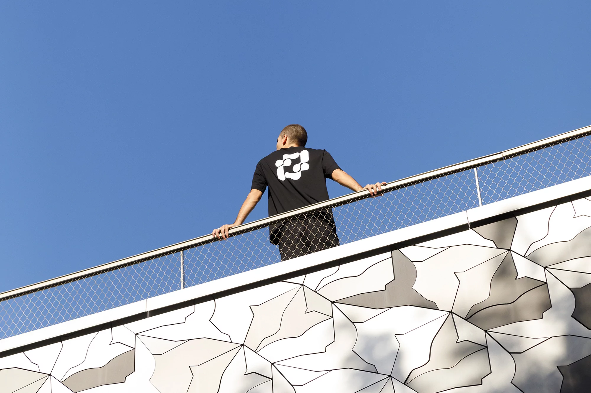 Adult male in black t-shirt at metal railing against blue sky and geometric gray and white patterned exterior wall.