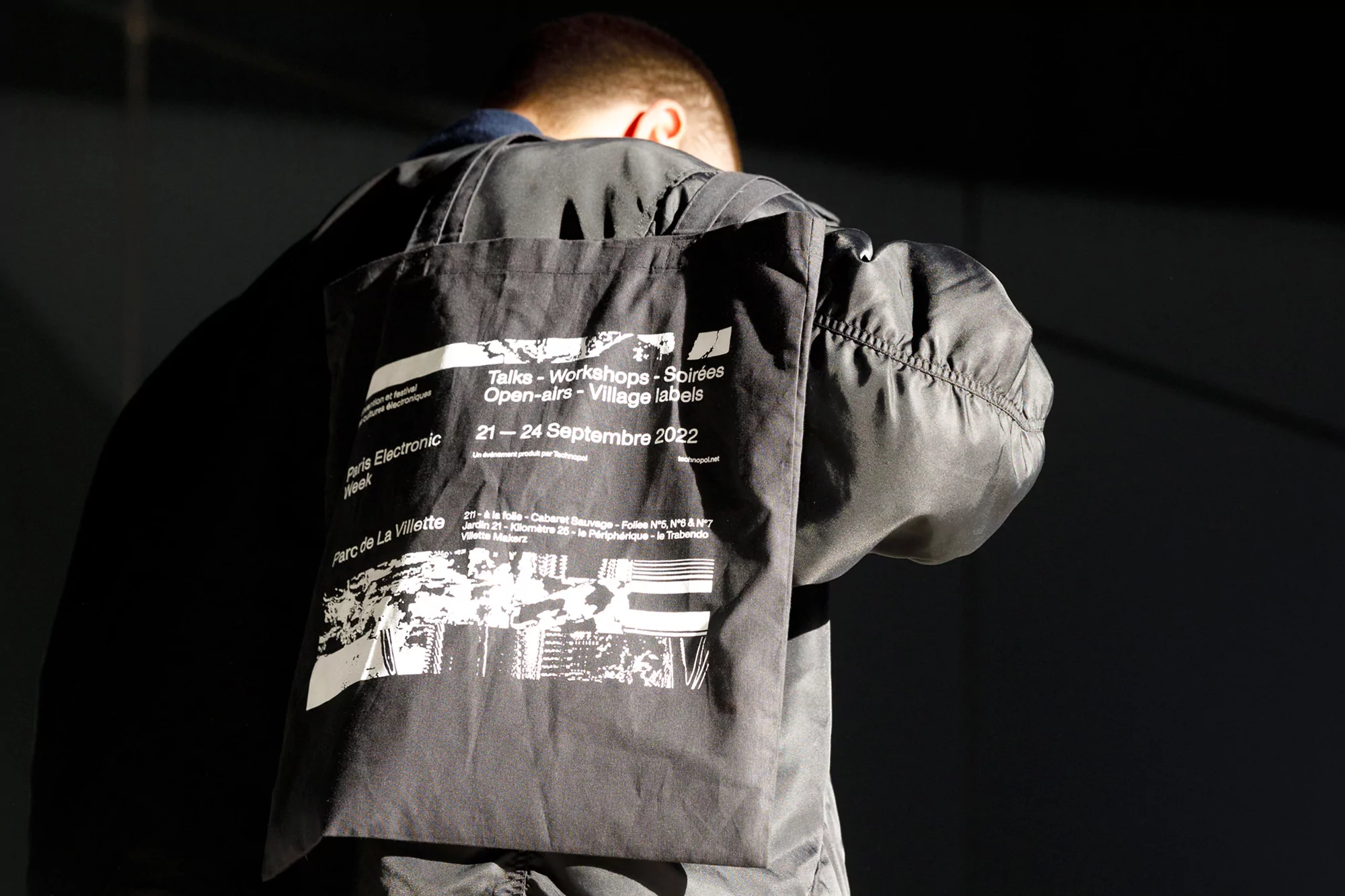 A person wears a black jacket with a tote bag printed with Paris Electronic Week event details against a dark background.