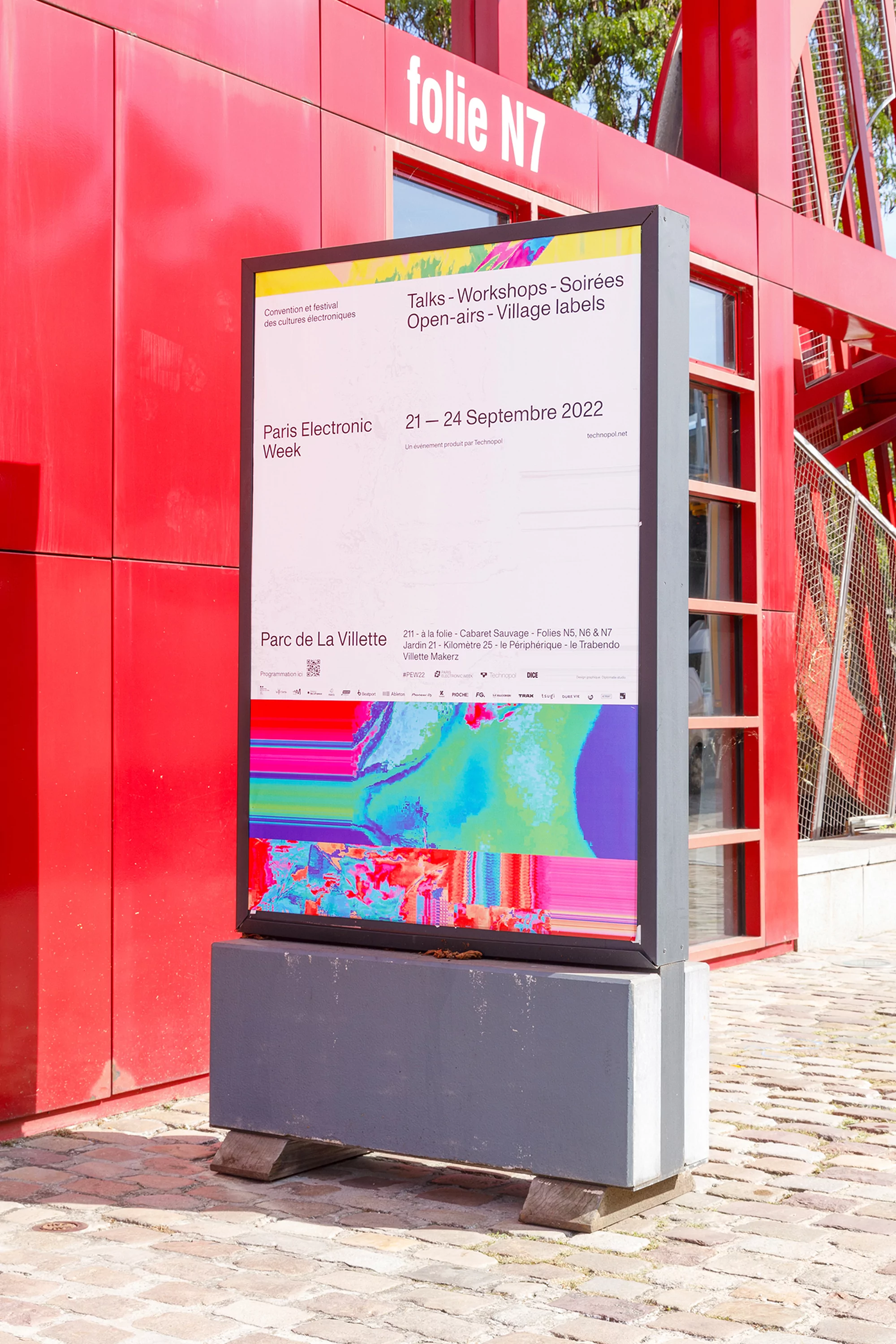 Outdoor digital signage displays Paris Electronic Week event poster near a modern red structure, showcasing talks and workshops in Parc de la Villette.