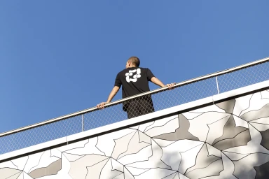Man from behind leaning on a metal fence overlooking a blue sky, wearing a black t-shirt with a white graphic design.