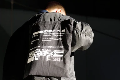 Person wearing a black bomber jacket carrying a black tote bag with white typographic print against a dark angled architectural backdrop.