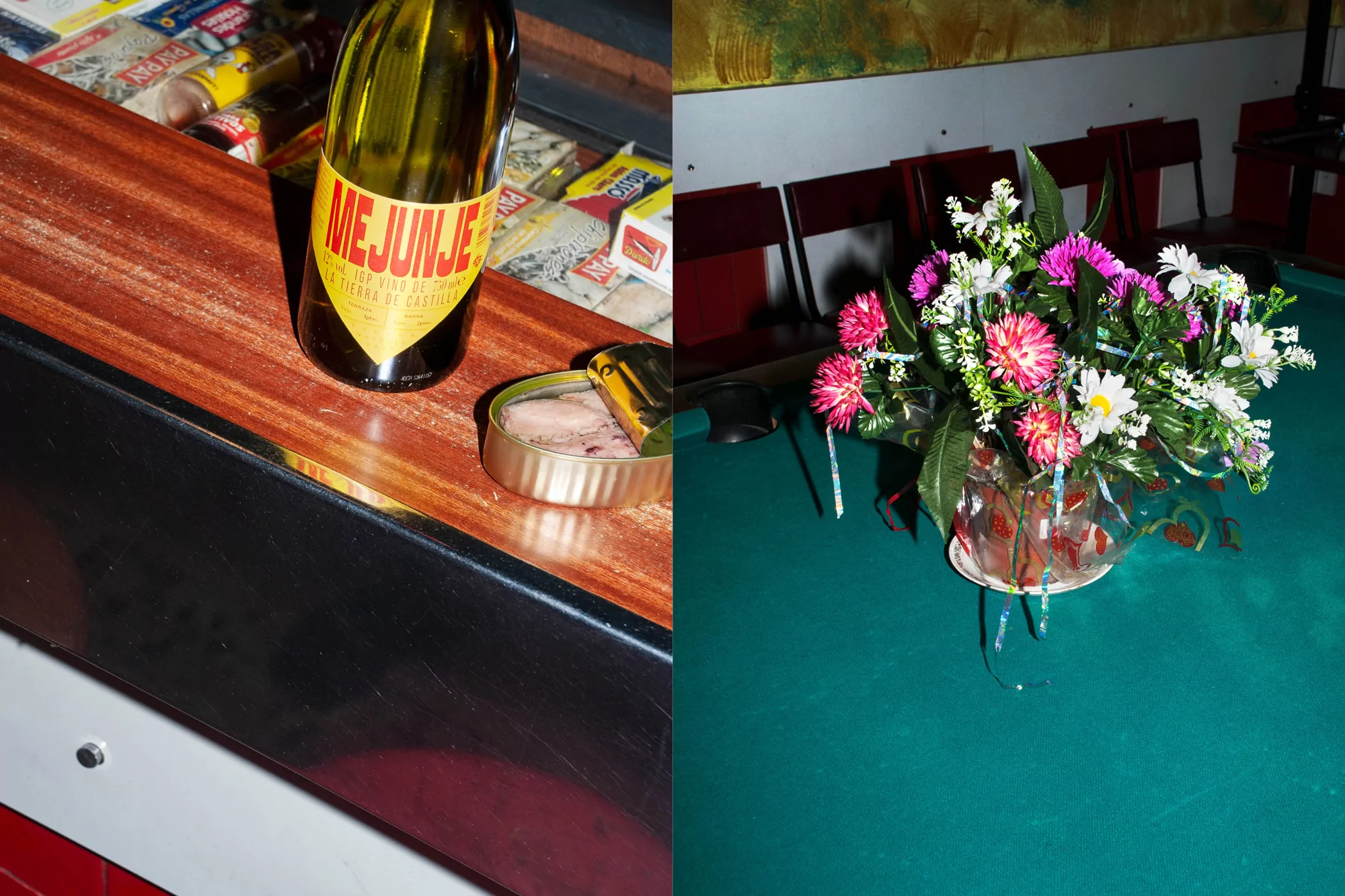 A bottle of wine and an open can of fish are set next to flowers on a pool table in a dimly lit indoor setting.