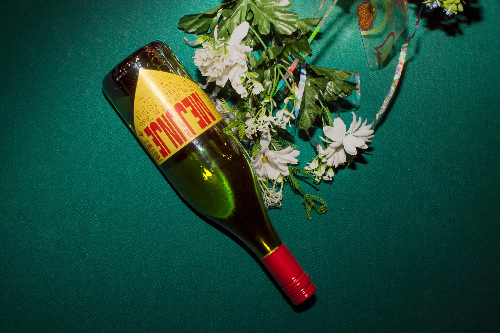 Wine bottle with red cap lays on its side, surrounded by white flowers on a green surface creating a celebratory or decorative still life.