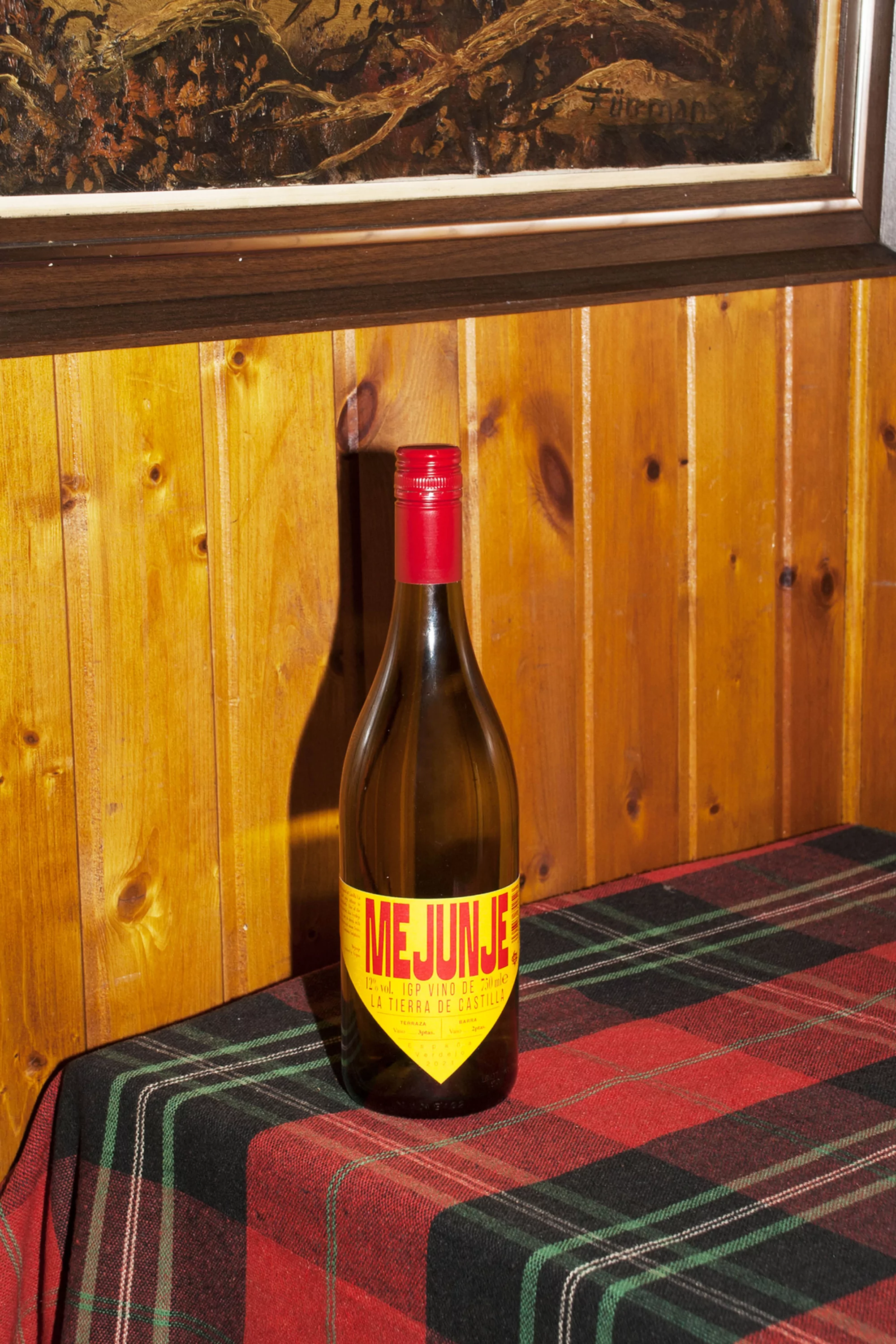 A bottle of Mejunje wine sits on a plaid tablecloth against a wood paneled wall with an old painting hanging above.