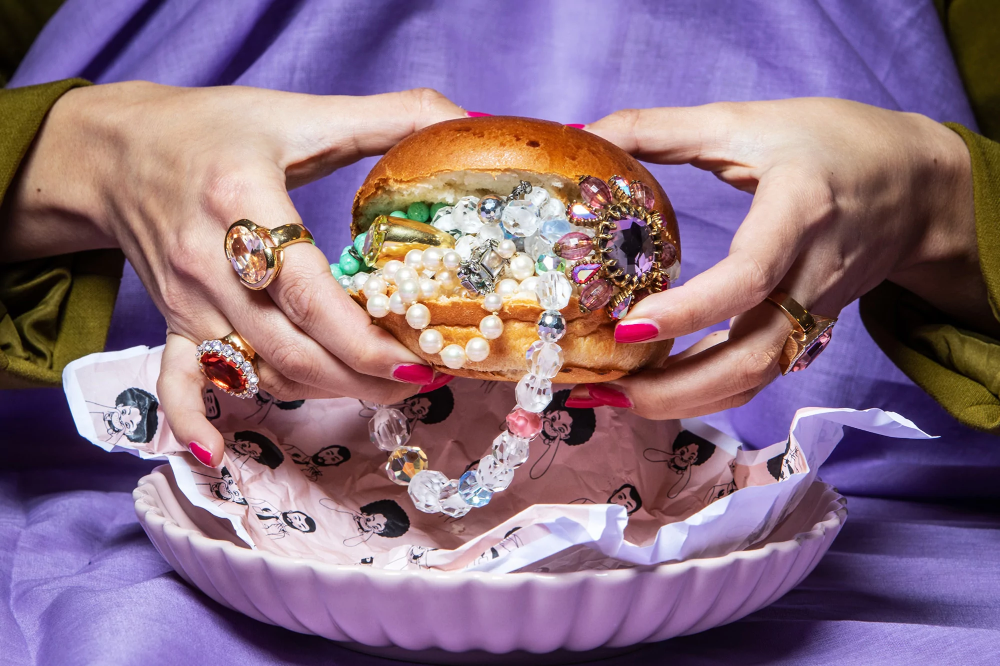 Close-up of hands holding a bun overflowing with jewelry, rings on fingers, in a patterned pink bowl, and set against a purple backdrop.