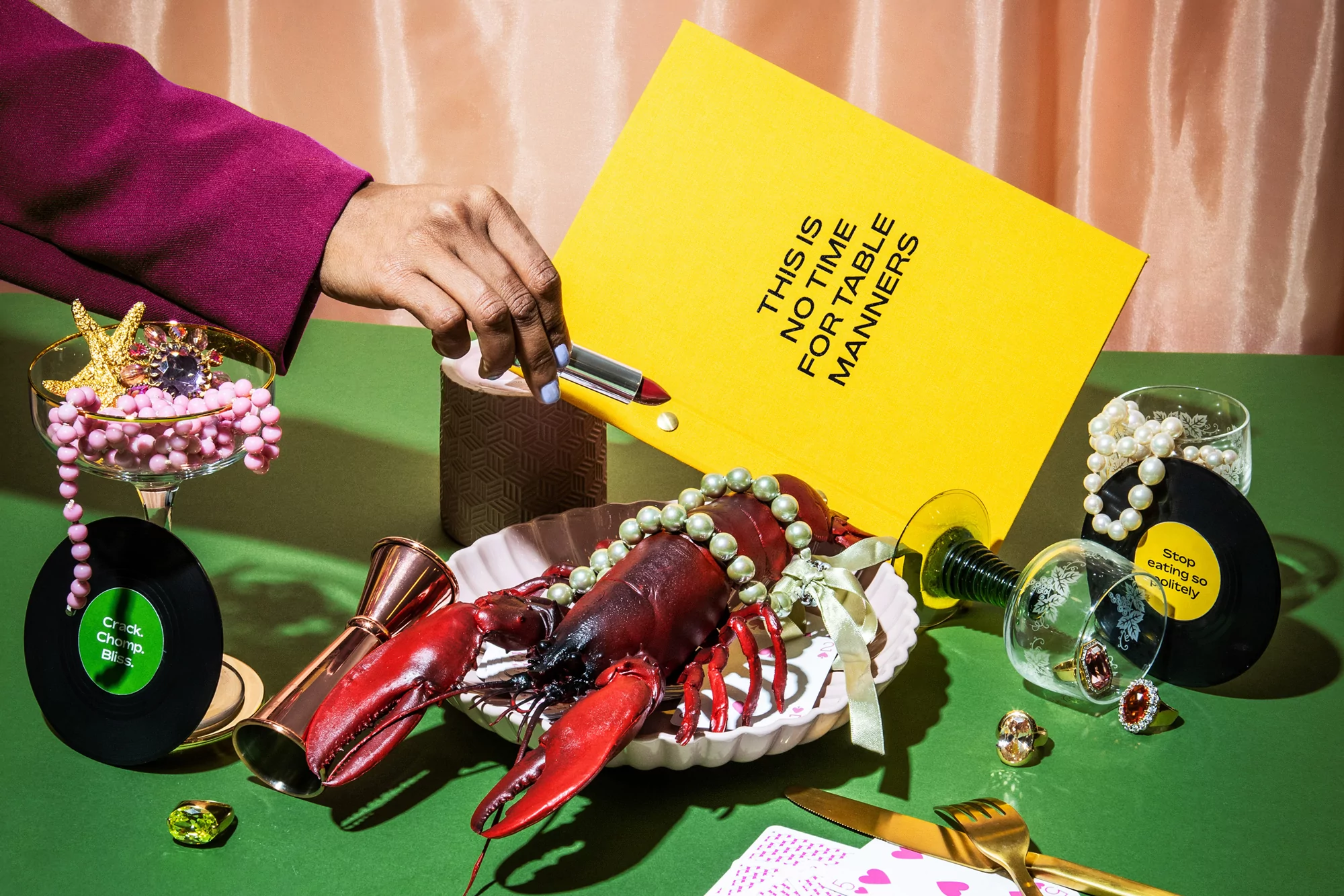 Still life featuring a lobster on a plate, surrounded by beads, jewelry, records, glassware, and a hand applying lipstick to a yellow card.
