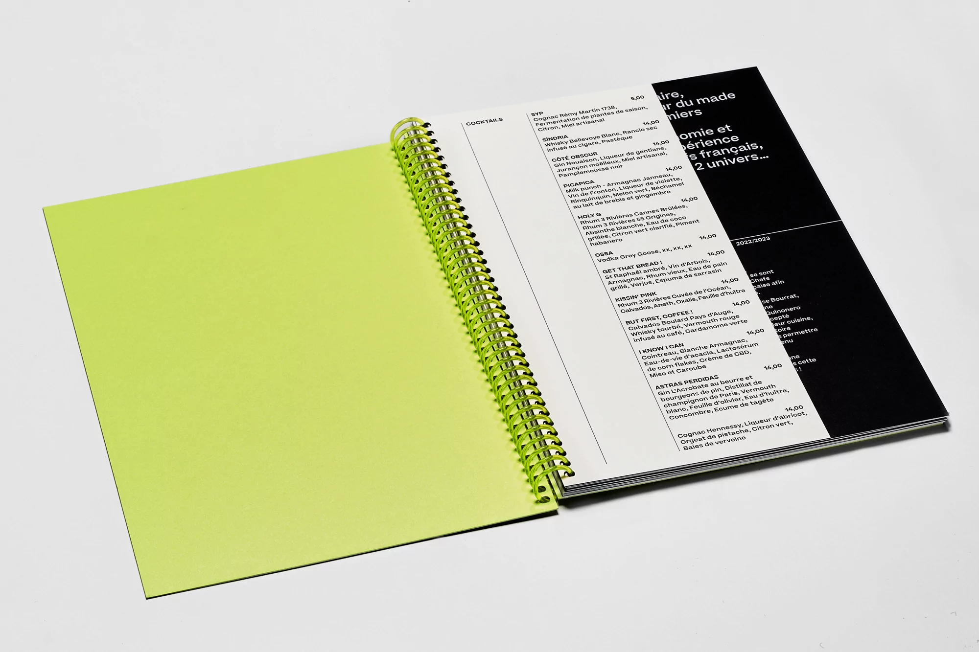 Open spiral-bound notebook with blank neon green page, facing a page of printed text, set against a plain white backdrop. Menu design.