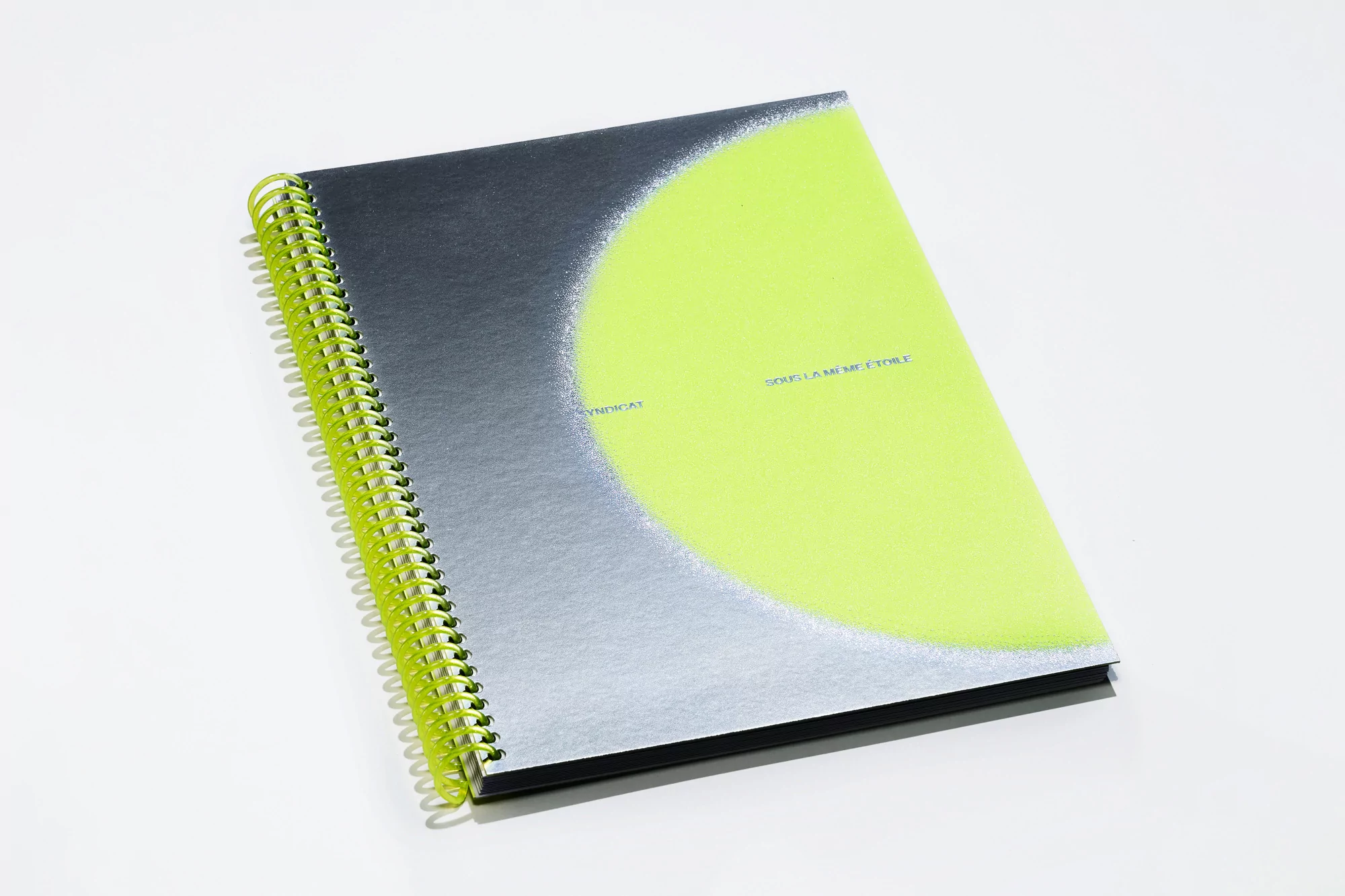 Eye-level close-up of a closed spiral notebook with a dark gray cover and a bright neon yellow circle on a solid white surface.