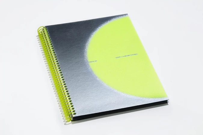 Eye-level close-up of a closed spiral notebook with a dark gray cover and a bright neon yellow circle on a solid white surface.
