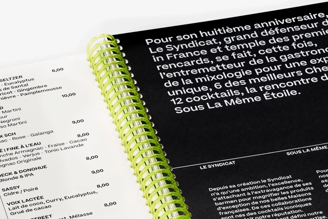 Close-up of a spiral-bound book featuring cocktail recipes in French, with a black and white color scheme, and a green spiral.