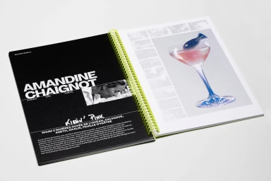 An open spiral-bound book on a white background, featuring a black page with bold white typography and a colorful cocktail photograph on the right.