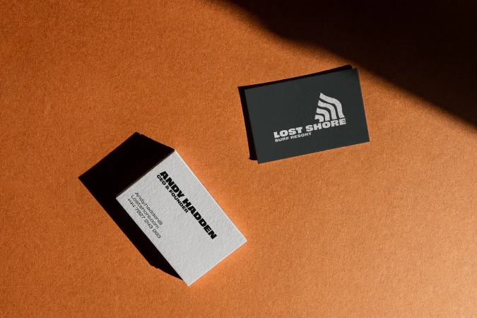 Two business cards, one white for Andy Hadden CEO & Founder, one black for Lost Shore Surf Resort, on a textured orange surface.