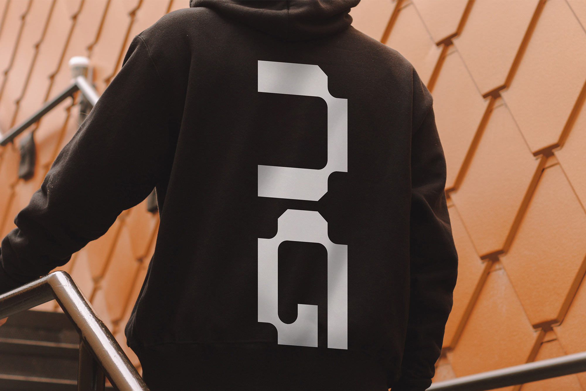 A person in a black hoodie with a large white design on the back stands near stairs with an orange textured wall in the background.