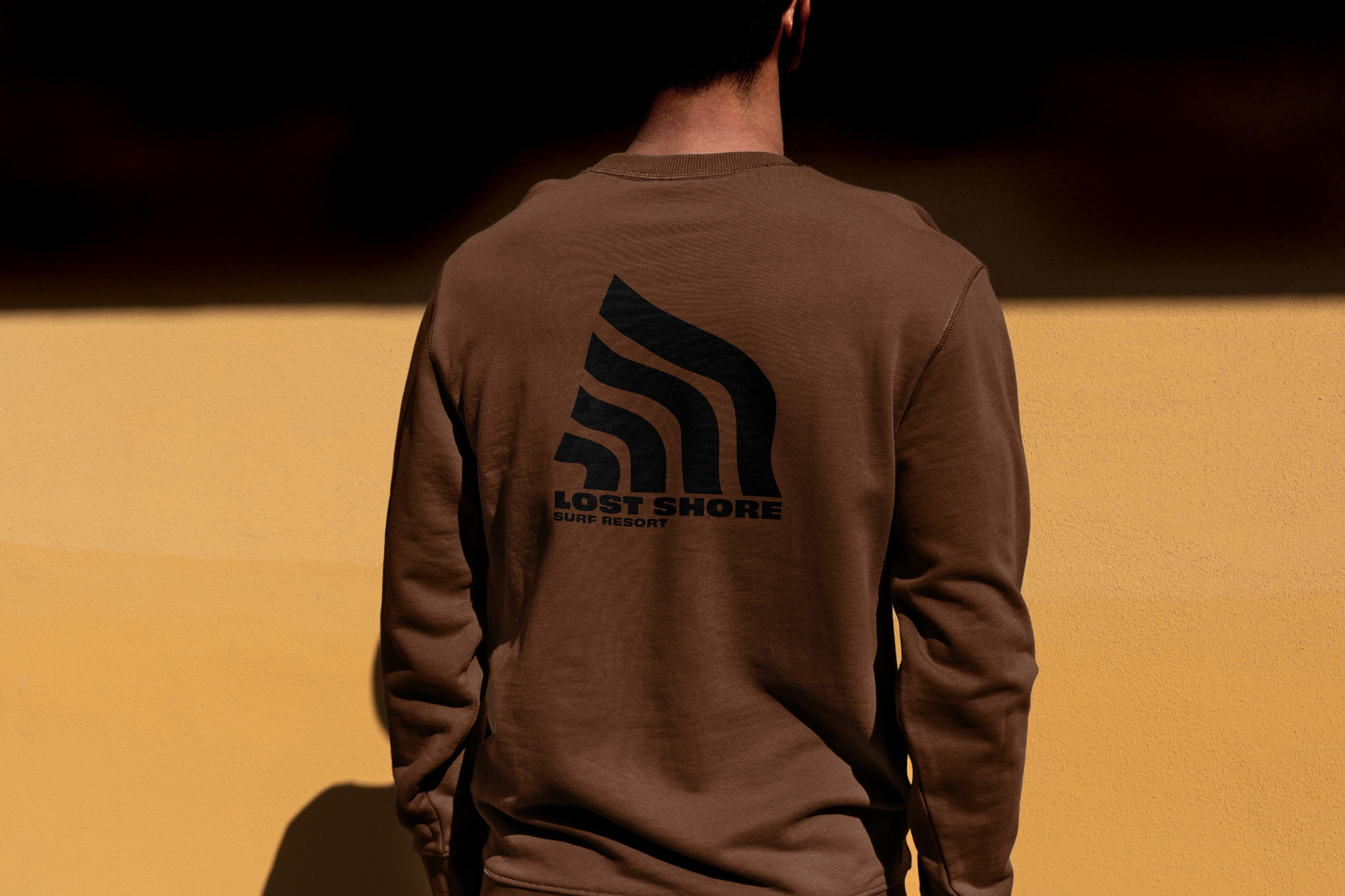 Person wearing brown sweatshirt with black logo, standing outdoors against a yellow wall in soft, natural light.