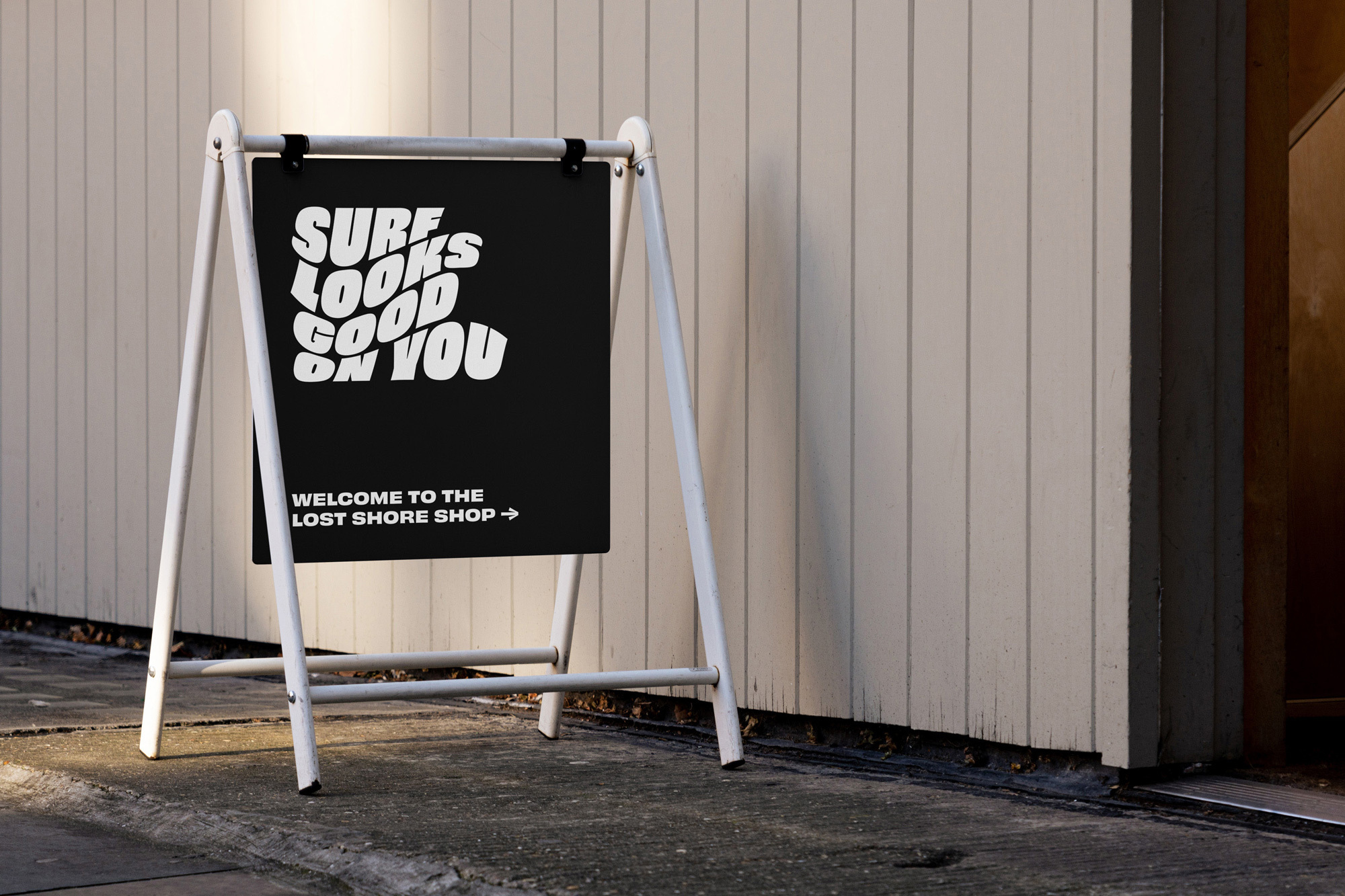 A-frame sandwich board on sidewalk displaying 'Surf Looks Good On You' advertising for The Lost Shore Shop near a white paneled wall.