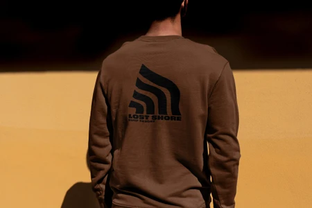 Rear view of a person wearing a brown sweatshirt with a black Lost Shore Surf Resort logo against a yellow wall