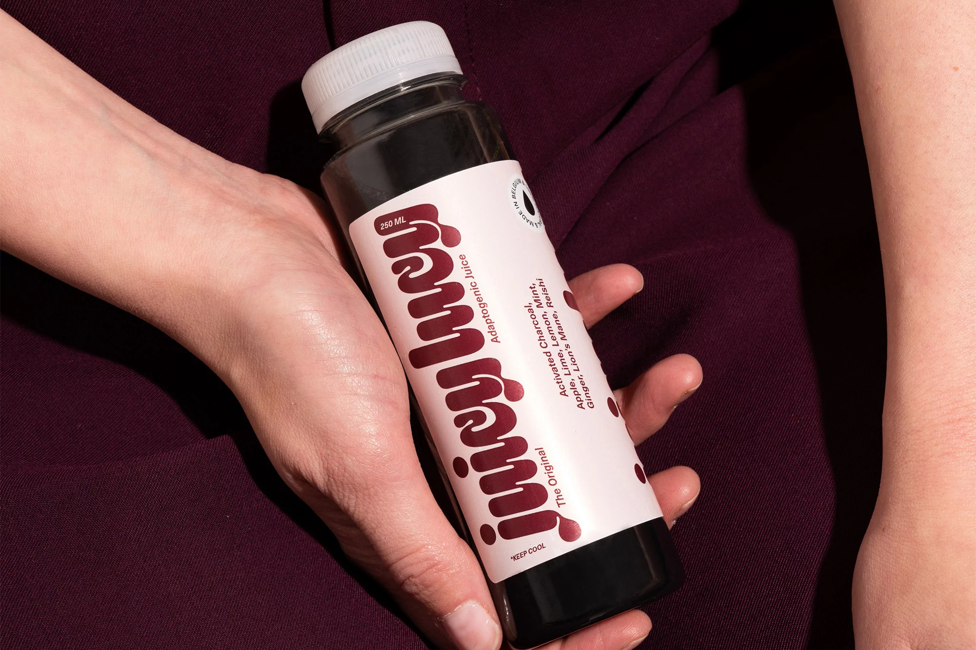 Adult hands hold a bottle of dark juice with a pink label, indoors on a purple fabric background. The label reads 'juicy lucy'.