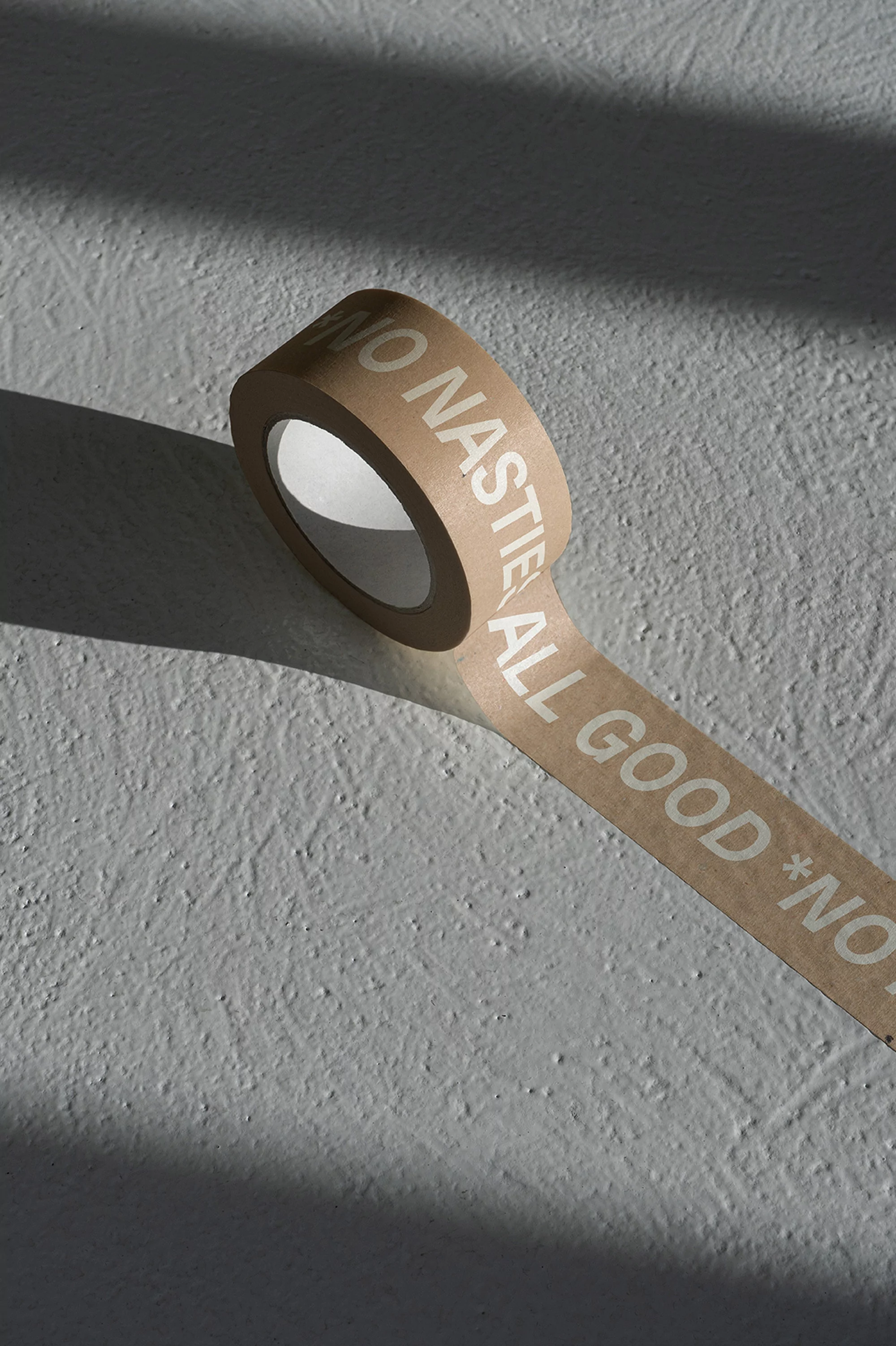 Roll of kraft tape with 'NO NASTIES ALL GOOD' text on a textured grey surface with cast shadows in a minimalist, indoor setting.