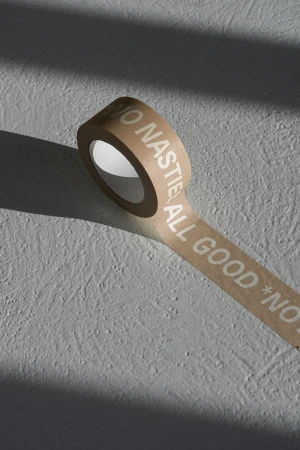 Roll of kraft tape with 'NO NASTIES ALL GOOD' text on a textured grey surface with cast shadows in a minimalist, indoor setting.