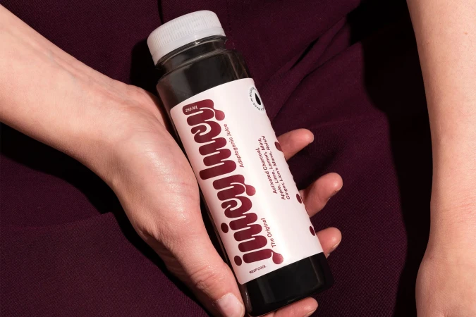 Close-up of hands holding a dark bottled juice with a white label for Juicy Lucy against a burgundy fabric background