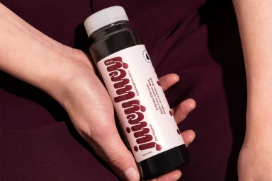 Close-up of hands holding a dark bottled juice with a white label for Juicy Lucy against a burgundy fabric background