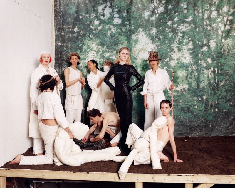 Fashion editorial shot of a group of models posing together on a dirt platform in a studio with painted forest background, wearing white and black clothing.