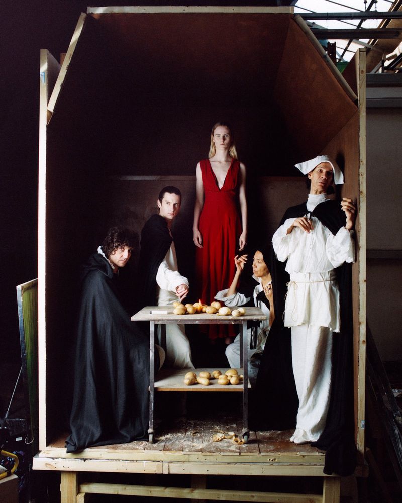 Five figures in classic garb staged inside a simple wooden structure, illuminated with a painterly light, evokes historical tableau vivant.