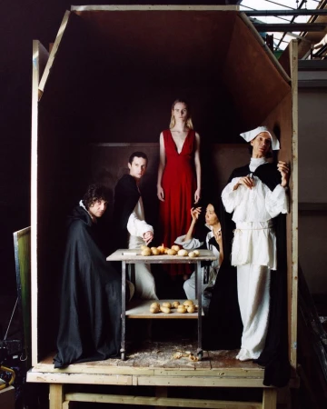 Five figures in classic garb staged inside a simple wooden structure, illuminated with a painterly light, evokes historical tableau vivant.