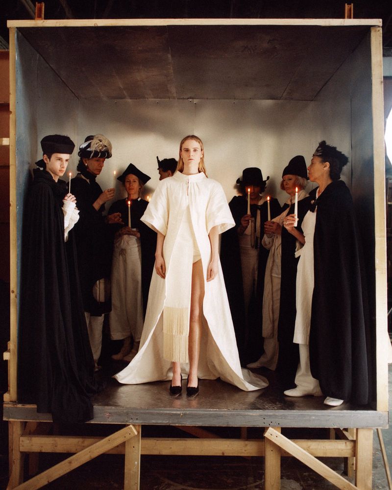 Fashion shoot in a box featuring a blonde woman in a white dress surrounded by people holding candles in a studio setting.