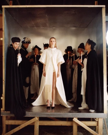 Fashion shoot in a box featuring a blonde woman in a white dress surrounded by people holding candles in a studio setting.