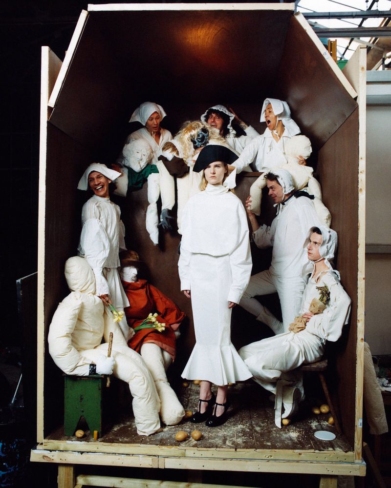 Artistic studio shot of a fashion model in a white dress surrounded by people and stuffed figures inside a wooden box-like structure.