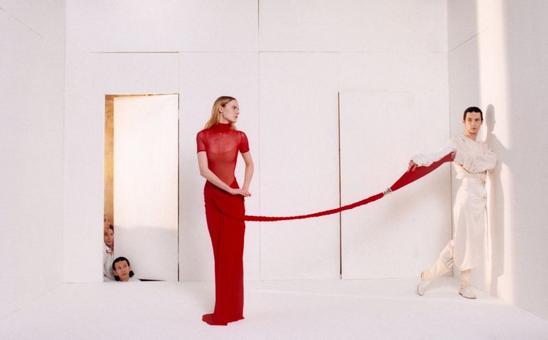 A woman in red dress holds a red rope that another person in white clothing is holding on to, posing in a white room.
